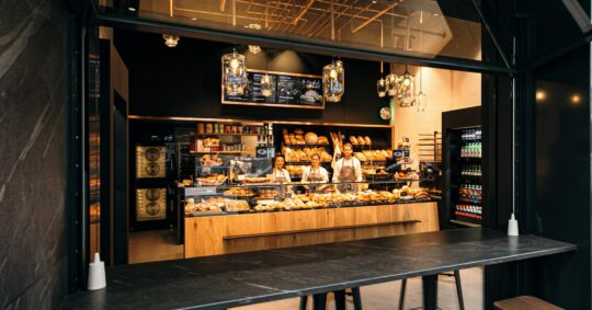 In einer modernen Bäckerei mit großen Glasfenstern wird eine Vielzahl von Brotsorten und Gebäck angeboten. Drei Mitarbeiter stehen hinter der Theke, bereit zu bedienen, und dahinter befinden sich Regale mit Backwaren und Getränken.