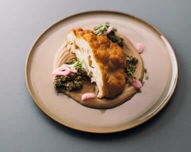 Ein Stück gebratener Blumenkohl liegt auf einem cremigen Püree, gekrönt von Mikrogrün und umgeben von einem Klecks rosa Sauce und kleinen Portionen einer fein gehackten Gemüsemischung auf einem beigen Teller.