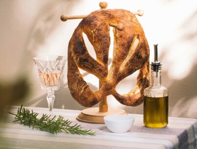Ein blattförmiger Brotlaib auf einem Holzständer neben einem Glas Roséwein, einem Rosmarinzweig, einer kleinen Schale und einer Flasche Olivenöl auf einem gestreiften Tischtuch.