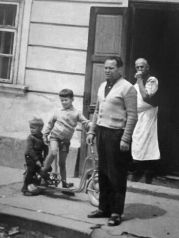 Ein Schwarz-Weiß-Foto von zwei Kindern und einem Mann, die vor einem Gebäude stehen. Ein weiterer Mann in einer Schürze steht in der Tür hinter ihnen und bedeckt seinen Mund teilweise mit der Hand. Ein Dreirad steht neben den Kindern.