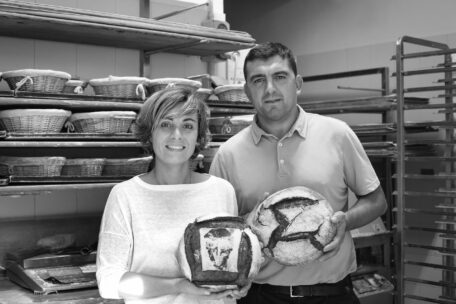 Eine Frau und ein Mann stehen in einer Bäckerei, lächeln und halten handwerklich gebackene Brote in der Hand. In den Regalen hinter ihnen stehen Körbe mit weiterem Brot. Das Bild ist in schwarz-weiß gehalten.