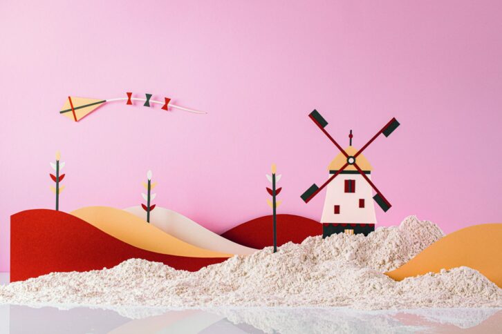 Eine Papierbastelszene zeigt eine Windmühle, Hügel und Blumen vor einem rosafarbenen Hintergrund, mit einem Drachen, der in den Himmel fliegt, und weißem, strukturiertem Material, das an Sand erinnert, im Vordergrund.