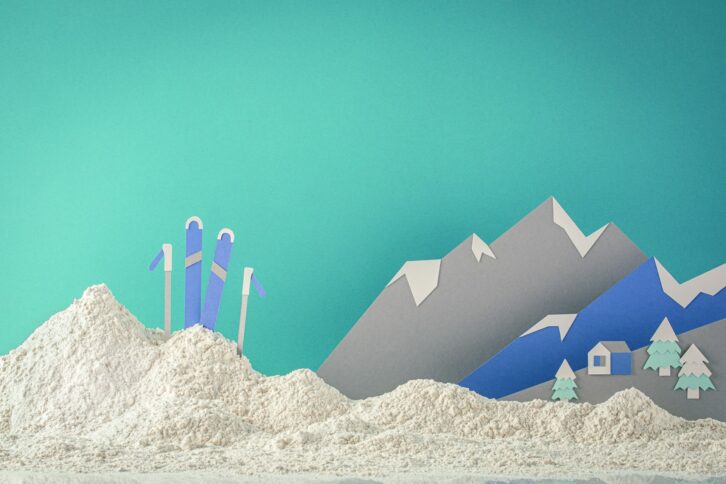 Scherenschnitt-Szene von verschneiten Bergen mit einer kleinen Hütte, Kiefern und drei Skiern mit Stöcken, die aus einem Schneehaufen herausragen, vor einem türkisfarbenen Hintergrund.
