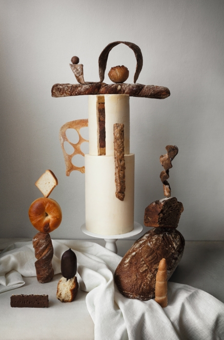 Ein künstlerisches Arrangement verschiedener Brote und Gebäckstücke, darunter Baguettes, Brote, Brötchen und eine Scheibe Brot, die auf und um einen hohen, weißen zylindrischen Kuchen auf einem Sockel vor einem neutralen Hintergrund balancieren.
