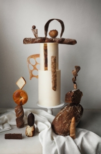 Ein kunstvolles Arrangement aus verschiedenen Brotsorten und Gebäck wird auf und um eine weiße zylindrische Torte balanciert, die auf einem Sockel steht, unter dem ein weißes Tuch drapiert ist, alles vor einem schlichten hellen Hintergrund.