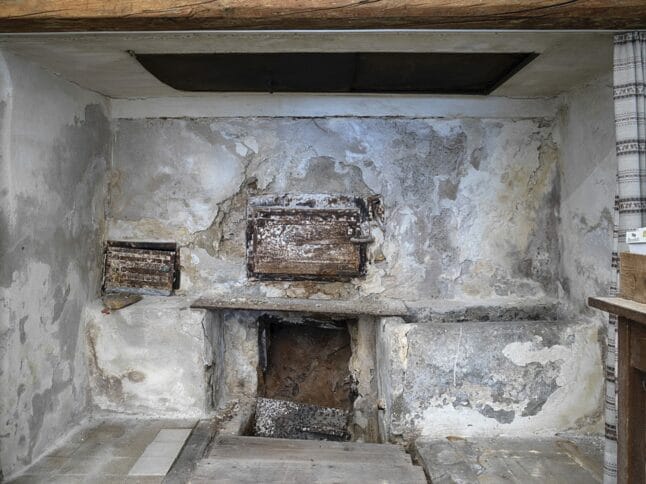 Ein alter, verwitterter Steinkamin mit zwei kleinen Metalltüren und einer rußverschmierten Öffnung vor einer rauen, verfärbten Wand in einem rustikalen Raum. Holzbalken und ein Teil eines gestreiften Vorhangs sind sichtbar.