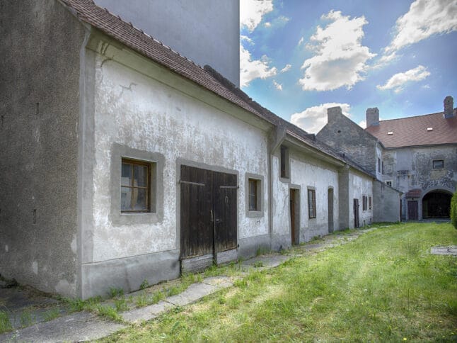 Eine Reihe alter, verwitterter Gebäude mit rissigen weißen Wänden und Holztüren steht neben einem grasbewachsenen Weg unter einem teilweise bewölkten Himmel. Die Szene hat eine rustikale, historische Atmosphäre.