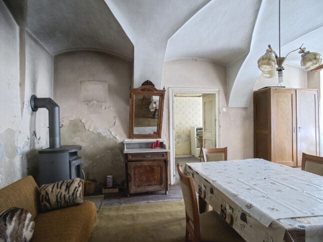 Ein rustikaler, leicht abgenutzter Speisesaal mit einem Tisch, der mit einer gemusterten Tischdecke bedeckt ist, einem hölzernen Schrank, einem alten Ofen, alten Möbeln und abblätternden Putzwänden unter einer gewölbten Decke.