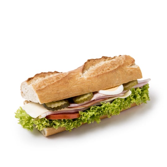 Ein Baguette-Sandwich, gefüllt mit Salat, Tomate, Schinken, Käse, Essiggurken und Scheiben von hartgekochtem Ei, auf weißem Hintergrund.
