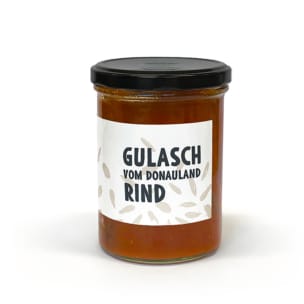 Ein Glasgefäß mit schwarzem Deckel, gefüllt mit rotbraunem Gulasch. Auf dem weißen Etikett steht in fetten schwarzen Buchstaben Gulasch vom Donauland Rind. Der Hintergrund ist einfarbig weiß.
