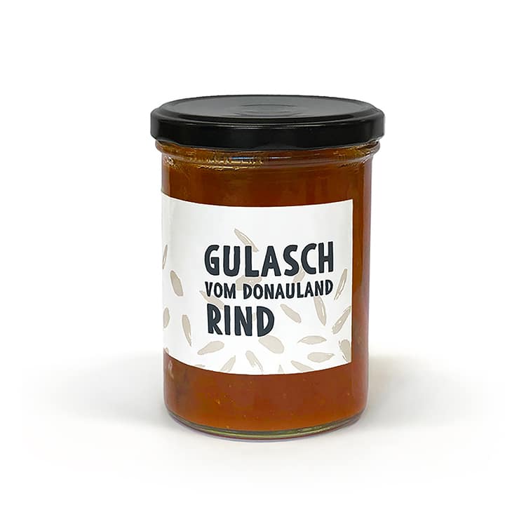 Ein Glasgefäß mit schwarzem Deckel, gefüllt mit rotbraunem Gulasch. Auf dem weißen Etikett steht in fetten schwarzen Buchstaben Gulasch vom Donauland Rind. Der Hintergrund ist einfarbig weiß.