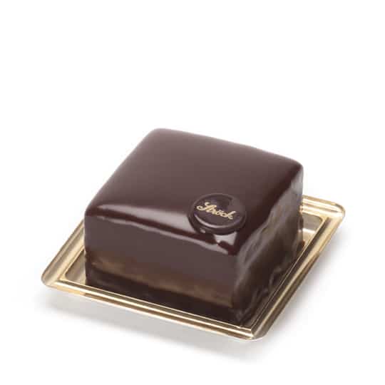 Eine quadratische Schokoladentorte mit glänzender Schokoladenglasur, gekrönt von einer kleinen runden Schokoladenscheibe mit dem Schriftzug Sacher, präsentiert auf einem goldenen Tablett.
