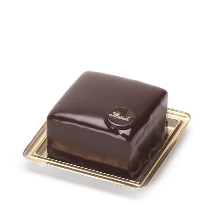 Eine quadratische Schokoladentorte mit glänzender Schokoladenglasur, gekrönt von einer kleinen runden Schokoladenscheibe mit dem Schriftzug Sacher, präsentiert auf einem goldenen Tablett.