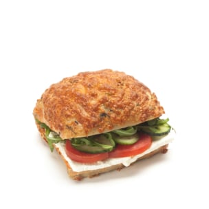 Ein Sandwich mit einem goldenen, käsigen Brötchen, gefüllt mit Tomatenscheiben, Gurken, Blattgemüse und einer Schicht weißem Käse, vor einem schlichten weißen Hintergrund.