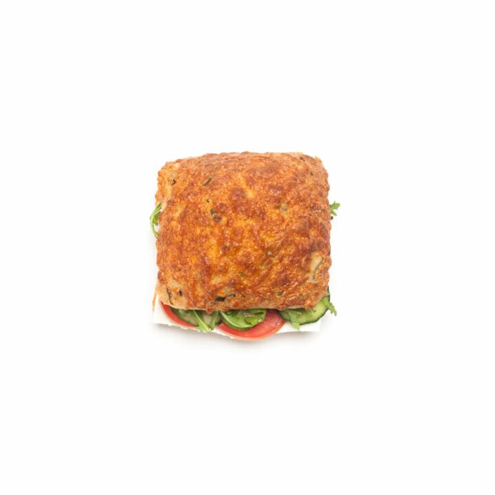 Ein quadratisches Sandwich mit einer goldenen, strukturierten Brotoberseite, gefüllt mit grünem Salat und roten Tomatenscheiben, zentriert auf einem weißen Hintergrund.