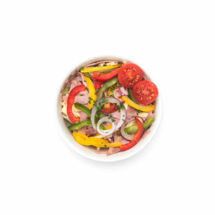Eine Schüssel Salat mit geschnittenen roten Zwiebeln, gelben und roten Paprikaschoten, Gurken, Kirschtomaten und Kräutern auf einer weißen Unterlage.
