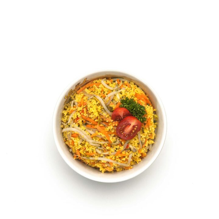 Eine Schüssel mit gelbem Reis und geraspelten Karotten, Bohnensprossen, halbierten Kirschtomaten und einem Zweig Petersilie auf weißem Hintergrund.