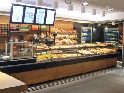 Eine moderne Bäckereiauslage mit Regalen, die mit verschiedenen Brotsorten, Gebäck und Backwaren gefüllt sind. Gekühlte Getränke stehen an der Rückwand, und über dem Kopf hängen digitale Bildschirme mit den Speisekarten. Der Bereich ist hell beleuchtet und sauber.