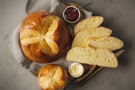 Ein runder Laib goldbraunes Brot mit einem Kreuzmuster, ein kleinerer ähnlicher Laib, drei Scheiben Brot, eine Schale mit Butter und eine Schale mit roter Marmelade, die auf einem Holzbrett mit einer Stoffserviette angeordnet sind.