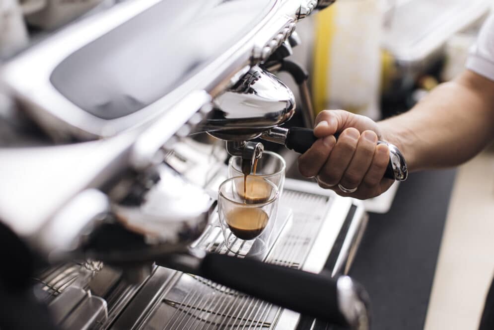Nahaufnahme einer Hand, die eine Espressomaschine bedient und zwei Espresso in Klarglastassen auf einem Metalltablett aufbrüht.