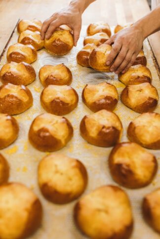 Die Hände einer Person, die frisch gebackene goldbraune Brötchen von einem mit Pergamentpapier ausgelegten Backblech auf einer hölzernen Oberfläche anordnet oder aufhebt.
