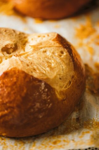 Eine Nahaufnahme eines goldbraunen, frisch gebackenen Brötchens mit gespaltener Oberseite, das auf Pergamentpapier liegt. Das Brot hat eine leicht glänzende Kruste und eine luftige Textur.
