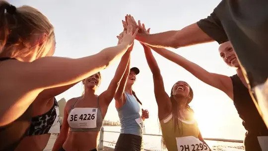 Eine Gruppe lächelnder Läuferinnen und Läufer mit Startnummern steht im Kreis und gibt sich zur Feier des Tages die Hand, während im Hintergrund die Sonne scheint.