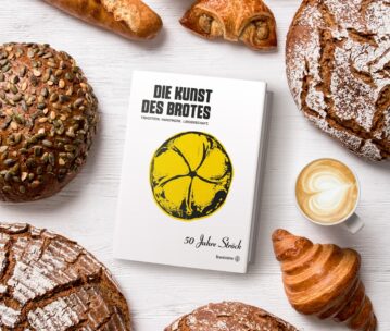 Ein Buch mit dem Titel Die Kunst des Brotes liegt auf einem weißen Tisch, umgeben von verschiedenen Broten, einem Croissant, einer Brezel und einer Tasse Milchkaffee mit herzförmigem Schaum. Das Buch ist mit einer gelben Brotillustration auf dem Einband versehen.