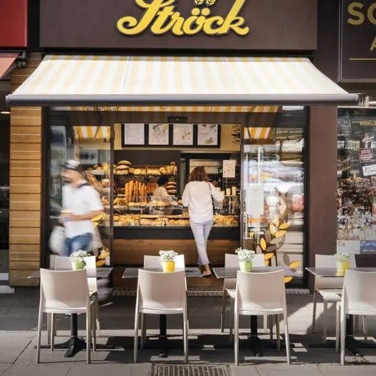 Außensitzbereich einer Bäckerei mit kleinen Tischen und Stühlen, jeder Tisch mit einem gelben Topf und weißen Blumen geschmückt. Zwei Personen sind in der Nähe der Auslage für Backwaren im Inneren des Ladens zu sehen, in der Backwaren ausgestellt sind.
