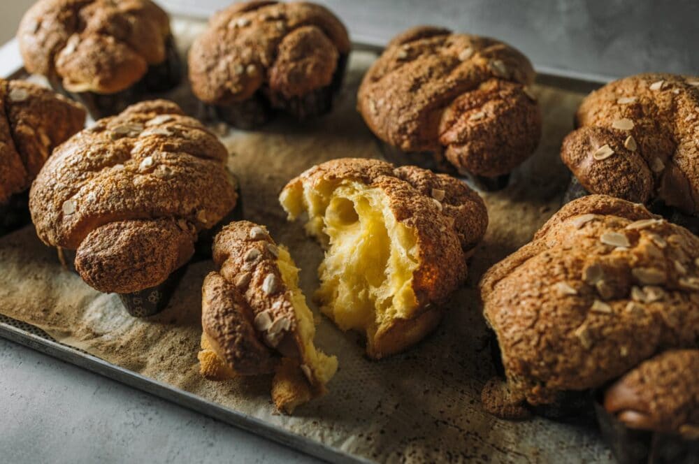 Auf einem mit Pergament ausgelegten Backblech liegen mehrere goldene, kuppelförmige Brioche-Brötchen mit knusprigen Oberseiten; ein Brötchen in der Mitte ist aufgerissen und zeigt sein weiches, luftiges und fluffiges Inneres.