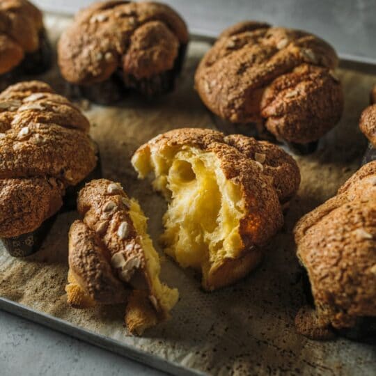 Auf einem mit Pergament ausgelegten Backblech liegen mehrere goldene, kuppelförmige Brioche-Brötchen mit knusprigen Oberseiten; ein Brötchen in der Mitte ist aufgerissen und zeigt sein weiches, luftiges und fluffiges Inneres.