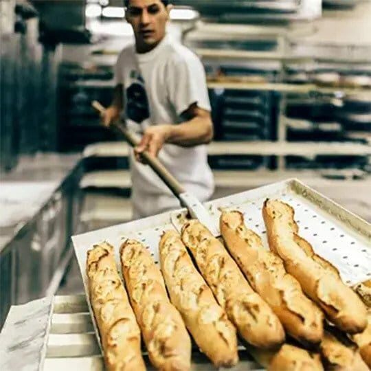Ein Bäcker im weißen Hemd nimmt mit einem langen Holzschäler frische Baguettes aus einem Industrieofen in einer Bäckerei, während im Hintergrund Tabletts und Regale zu sehen sind.