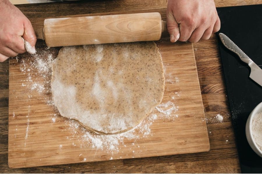 Eine Person rollt den Teig mit einem hölzernen Nudelholz auf einem bemehlten Holzbrett aus. Ein Messer und eine kleine Schüssel mit Mehl stehen auf einer schwarzen Matte daneben.