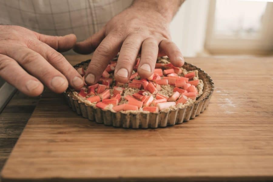 Nahaufnahme der Hände einer Person, die gehackte Rhabarberstücke auf einer ungebackenen Torte in einer geriffelten Form auf einem hölzernen Schneidebrett anordnet. Die Szene erinnert an das Backen in einer Küche.