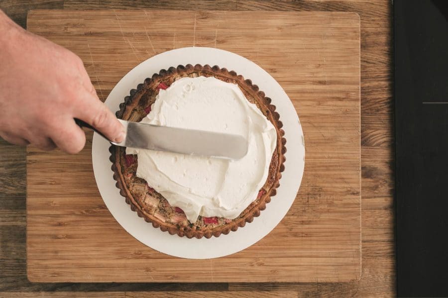 Eine Person streicht mit einem Spatel weißen Zuckerguss auf eine runde Torte, die auf einem weißen Teller auf einem hölzernen Schneidebrett liegt.