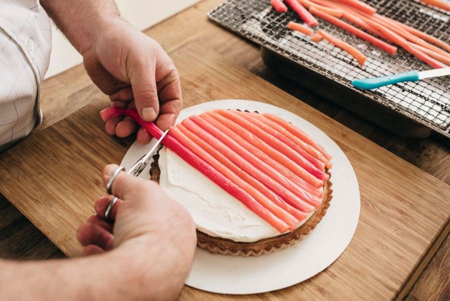 Eine Person legt dünne, rosa Rhabarberstreifen auf eine mit weißer Creme bestrichene Torte und schneidet die Enden mit einer Schere ab. Ein Tablett mit weiteren Rhabarberstreifen steht in der Nähe auf einer hölzernen Arbeitsplatte.
