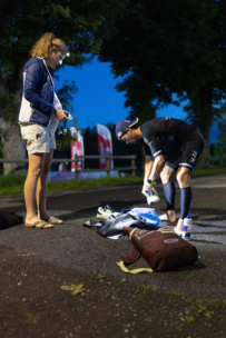 Ein Mann in einem Neoprenanzug bereitet in der Morgendämmerung die Ausrüstung vor, während eine Frau in Jacke und Shorts in der Nähe steht und die Ausrüstung hält. Im Hintergrund sind Bäume und rote Fahnen zu sehen.