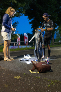 Ein Mann in einem Triathlonanzug bereitet die Ausrüstung auf dem Boden vor, einschließlich der Schuhe und des Neoprenanzugs, während eine Frau in Freizeitkleidung ihm am frühen Morgen oder am späten Abend im Freien assistiert.