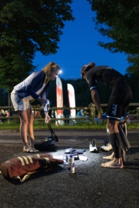 Zwei Personen stehen in der Morgen- oder Abenddämmerung auf einer Straße, beleuchtet von einer Stirnlampe. Der eine trägt Sportkleidung und ist barfuß, der andere hilft, vor einem Rennen Gegenstände auf dem Boden zu sortieren, darunter Schuhe und Flaschen. Im Hintergrund sind Bäume und Fahnen zu sehen.
