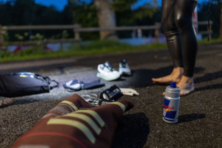 Eine Person in einem Neoprenanzug steht barfuß auf einer nassen Straße in der Abenddämmerung, in der Nähe von Laufschuhen, Handschuhen, einer Red Bull Trinkflasche und einem Rucksack. Die Szene scheint Teil einer Sportveranstaltung im Freien oder eines Triathlon-Übergangs zu sein.