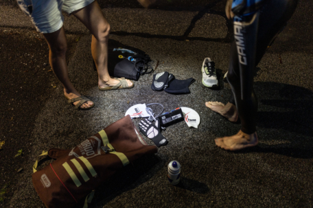 Zwei Personen stehen nachts barfuß auf dem Bürgersteig, in der Nähe von Triathlonausrüstungen wie Schwimmflügeln, Badekappen, einem Neoprenanzug, einer gestreiften Tasche und einer mit einer Taschenlampe beleuchteten Wasserflasche.