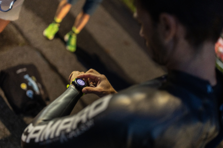 Eine Person in einem Neoprenanzug überprüft eine Smartwatch, wobei der Fokus auf ihrem Handgelenk liegt und der unscharfe Hintergrund eine andere Person zeigt, die in der Nähe steht. Der Boden ist gepflastert und es liegen Sportgeräte auf dem Boden.