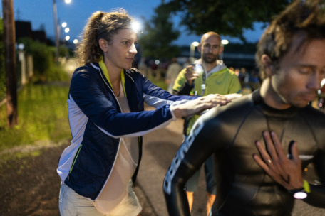 Eine Frau mit Stirnlampe und Sportjacke massiert die Schultern einer Person in einem Neoprenanzug im Freien in der Abenddämmerung. Eine andere Person steht im Hintergrund und hält einen Becher.