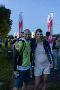 Zwei lächelnde, sportlich gekleidete Personen posieren in der Abenddämmerung zusammen im Freien. Eine Person ist sichtlich schwanger. Im Hintergrund sind rote Veranstaltungsbanner und andere Teilnehmer zu sehen.