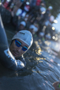 Ein Triathlet mit Badekappe und blauer Schwimmbrille schwimmt im Wasser und hält sich am Rand fest, während andere Athleten und Zuschauer im Hintergrund verschwommen zu sehen sind.