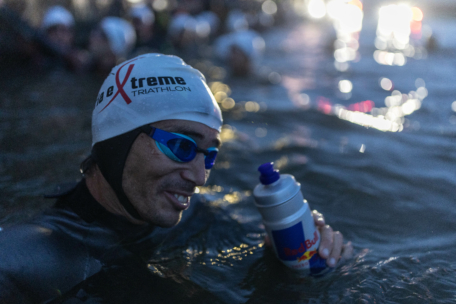 Ein Triathlet mit weißer Badekappe und blauer Schwimmbrille lächelt, während er eine Red Bull-Wasserflasche im Wasser hält, umgeben von anderen Schwimmern in Neoprenanzügen, mit hellen Lichtern im Hintergrund.