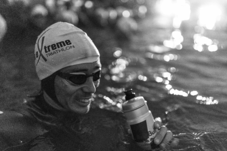 Ein Triathlet mit Badekappe und Schwimmbrille lächelt, während er eine Wasserflasche im offenen Wasser hält, umgeben von anderen Schwimmern, mit hellem Licht im Hintergrund.