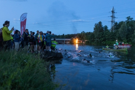 Eine Gruppe von Schwimmern in Neoprenanzügen wetteifert in der Abenddämmerung in einem Fluss, während die Menschen am Ufer jubeln und Fotos machen; im Hintergrund sind ein Boot mit Lichtern und ein Feuer in der Nähe einer Brücke zu sehen.