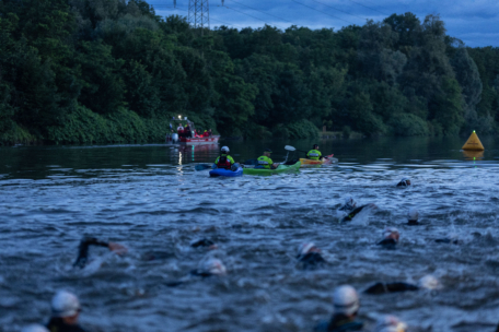 Eine Gruppe von Schwimmern nimmt an einem Freiwasserrennen teil, das von Kajakfahrern und einem Rettungsboot genau beobachtet wird, mit bewaldeten Flussufern und einer gelben Boje im Hintergrund.