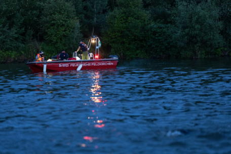 Ein rotes Rettungsboot mit der Aufschrift "FEUERWEHR FELDKIRCHEN" liegt in der Abenddämmerung auf einem See in der Nähe eines bewaldeten Ufers. An Bord befinden sich mehrere Personen in Schutzausrüstung, die von einem hellen Licht auf dem Boot beleuchtet werden.